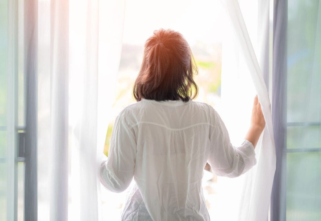 A person with dark hair, wearing a white shirt, stands facing a bright window, holding open white curtains to welcome the morning light during daylight saving time.