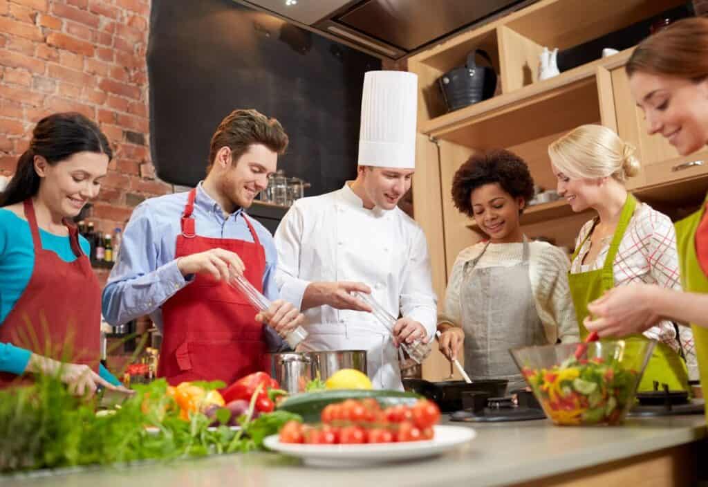 A chef instructs a group of people wearing aprons as they cook together in a kitchen, with fresh vegetables on the counter.